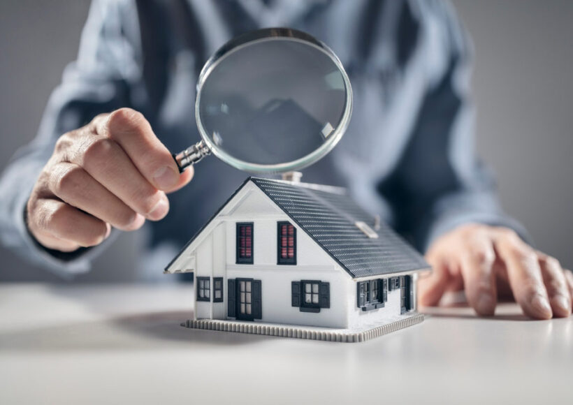 A man holds a magnifying glass up to a model home, symbolizing a pre-listing inspection.
